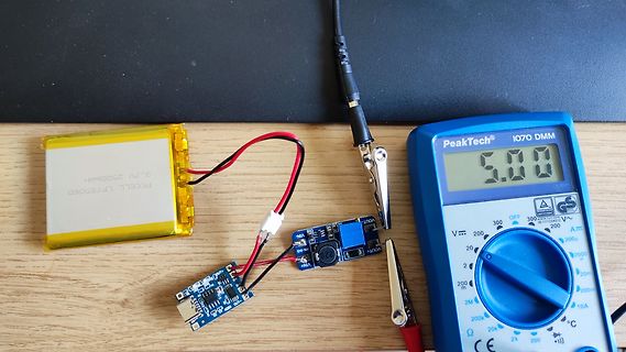 LiPo-Akku, Lademodul, Mikrocontroller und Multimeter auf Holzuntergrund
